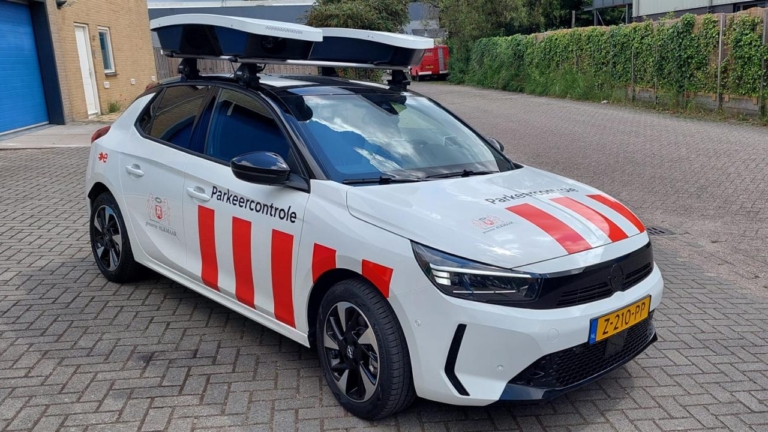 Witte parkeercontroleauto met rood-witte strepen en camera’s op het dak, geparkeerd op een klinkerweg.