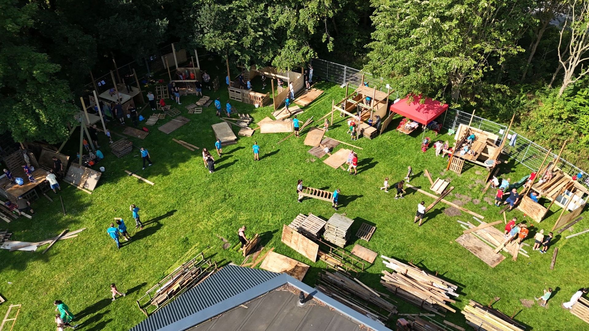 Bovenaanzicht van kinderen die houten hutten bouwen in een grasveld omringd door bomen.