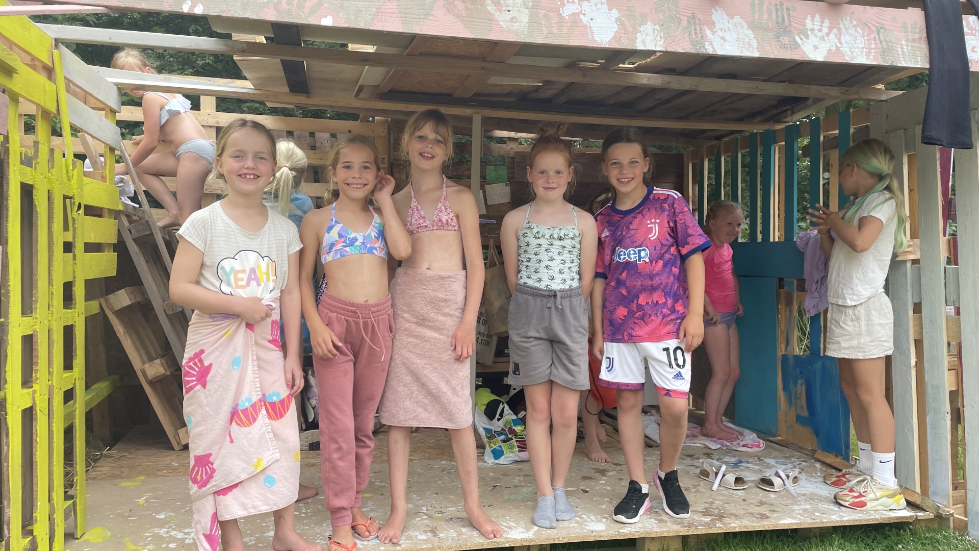 Groep kinderen die lachen en poseren in een zelfgebouwd houten fort, gekleed in zomerkleding.