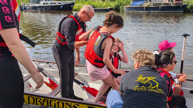 Mensen helpend kinderen in een boot met rode reddingsvesten, dicht bij een waterkant.