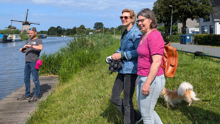 Twee vrouwen en een man met een camera wandelen langs een kanaal met een molen op de achtergrond, begeleid door een kleine hond.