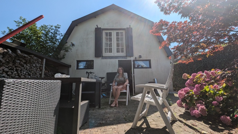 Vrouw zittend op een tuinstoel in een tuin met een wit huis op de achtergrond, omringd door een fiets, bloemen en tuinmeubels.