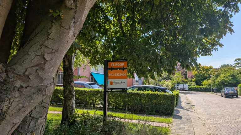 Straatbeeld met grote boom en "Te Koop" bord van Kuypers en Blom makelaars.