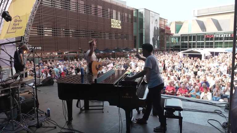 Twee muzikanten op een podium kijken naar een groot publiek buiten voor een gebouw met de tekst "Stedelijk Museum Alkmaar".