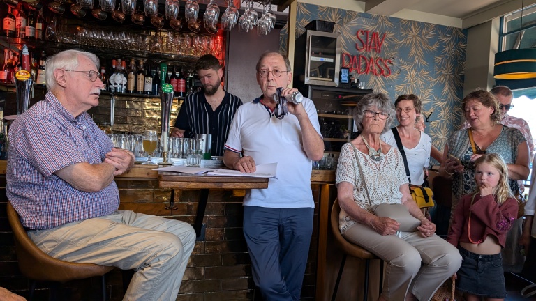 Mensen verzamelen zich in een bar terwijl een man in een microfoon spreekt;  Op de achtergrond staat een neonbord met de tekst "BLIJF RADAS".