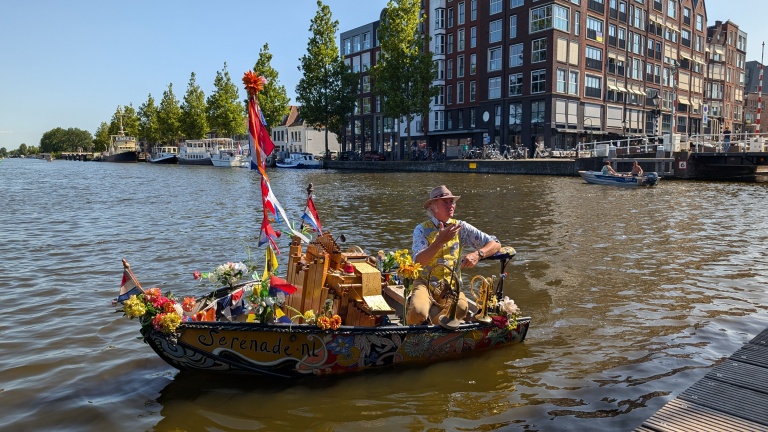 Man in versierde boot met muziekinstrumenten op kanaal, gebouwen en boten op de achtergrond.