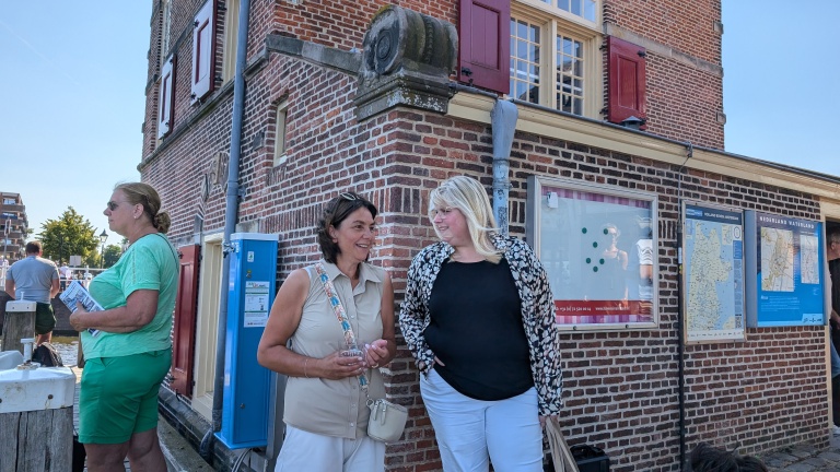 Twee vrouwen praten naast een bakstenen gebouw met informatiepanelen en een blauwe paal.