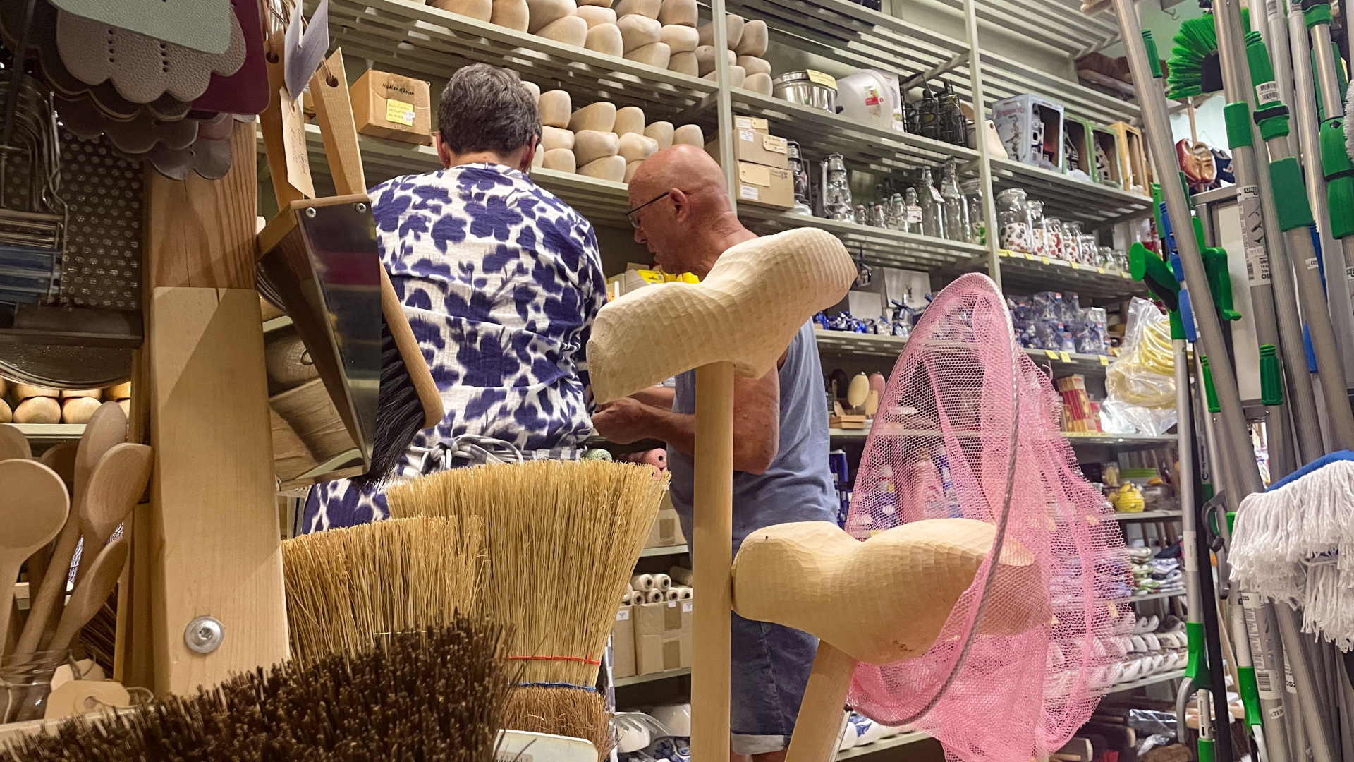 Mensen winkelen in een winkel vol huishoudelijke artikelen zoals bezems, borstels en andere schoonmaakspullen.