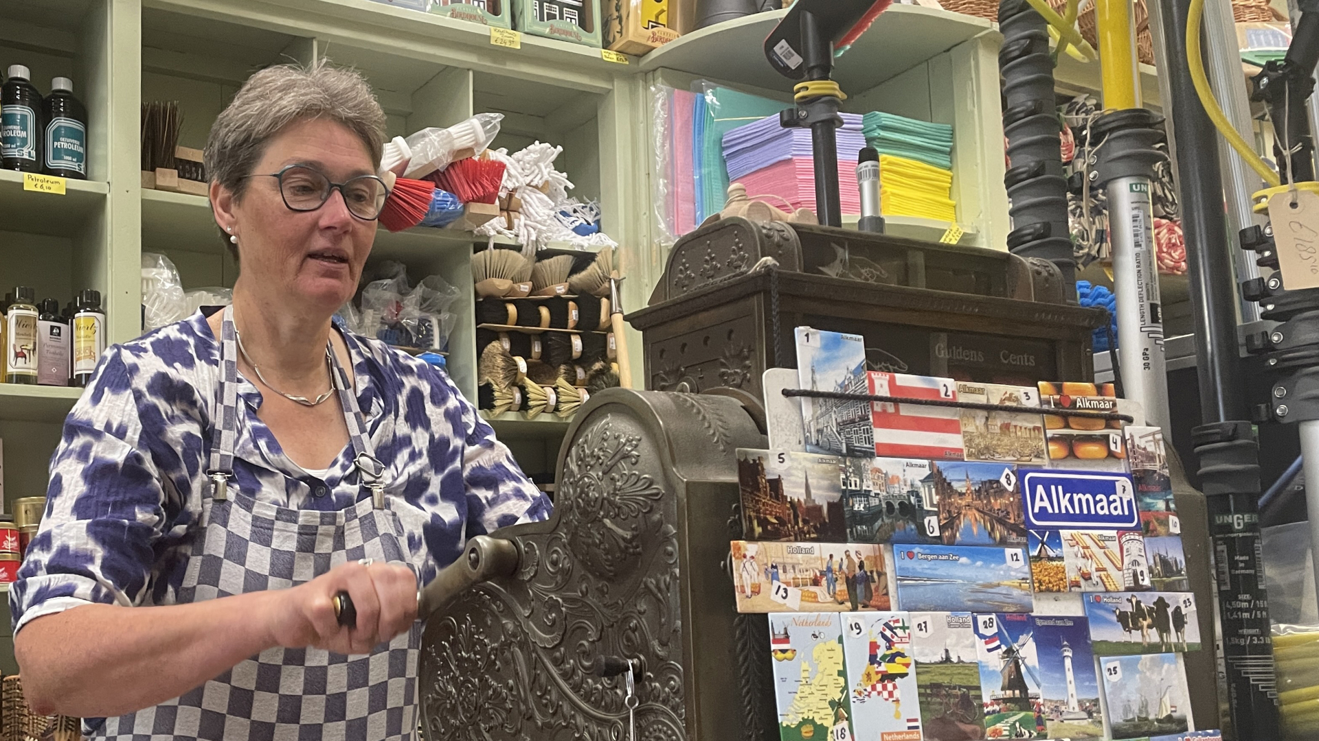 Een vrouw in een winkel met huishoudelijke artikelen en schoonmaakbenodigdheden die achter een antieke kassa staat, met ansichtkaarten van Alkmaar op de voorgrond.