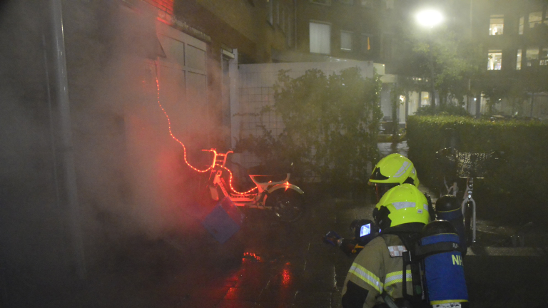 Brandweerlieden blussen een brand bij een fiets met rode lichtjes in een nachtelijke straat.