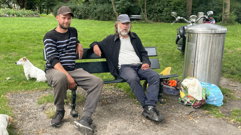 Twee mannen zitten op een bankje in een park met een hond en enkele tassen naast een prullenbak.