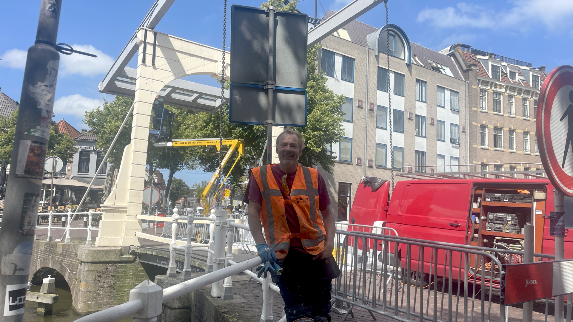 Bouwvakker met oranje veiligheidshesje leunt tegen een hek bij een brug in een stadscentrum met rode bestelwagens en een restaurant op de achtergrond.