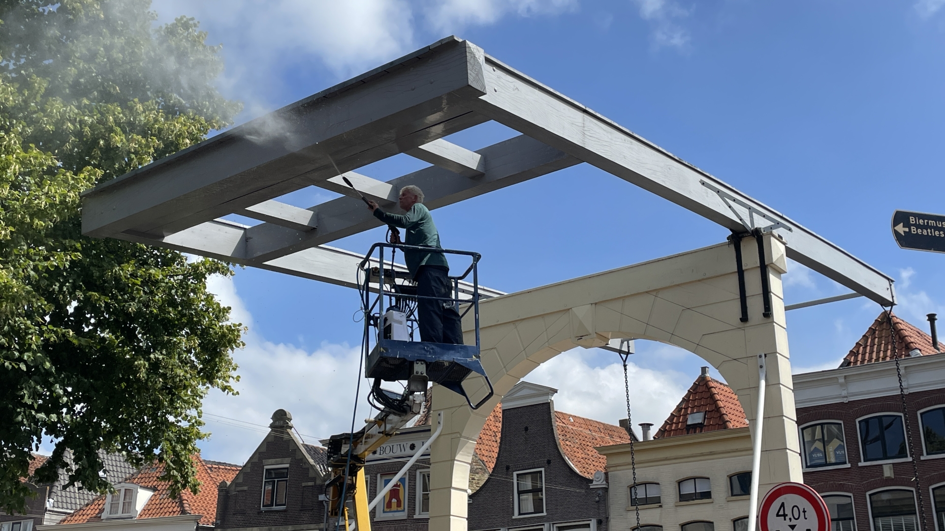 Man op hoogwerker schildert een wit houten structuur in een stadsomgeving.