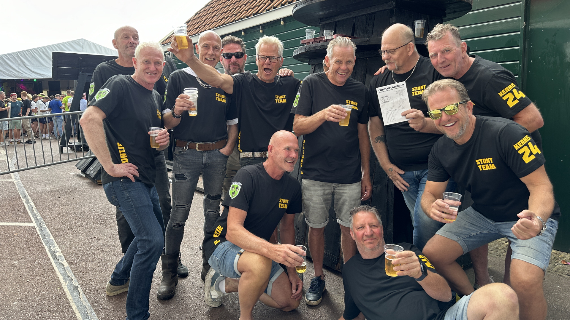 Een groep mannen in zwarte T-shirts met opschrift "Stunt Team" bij een buitenevenement, lachend en bier drinkend.