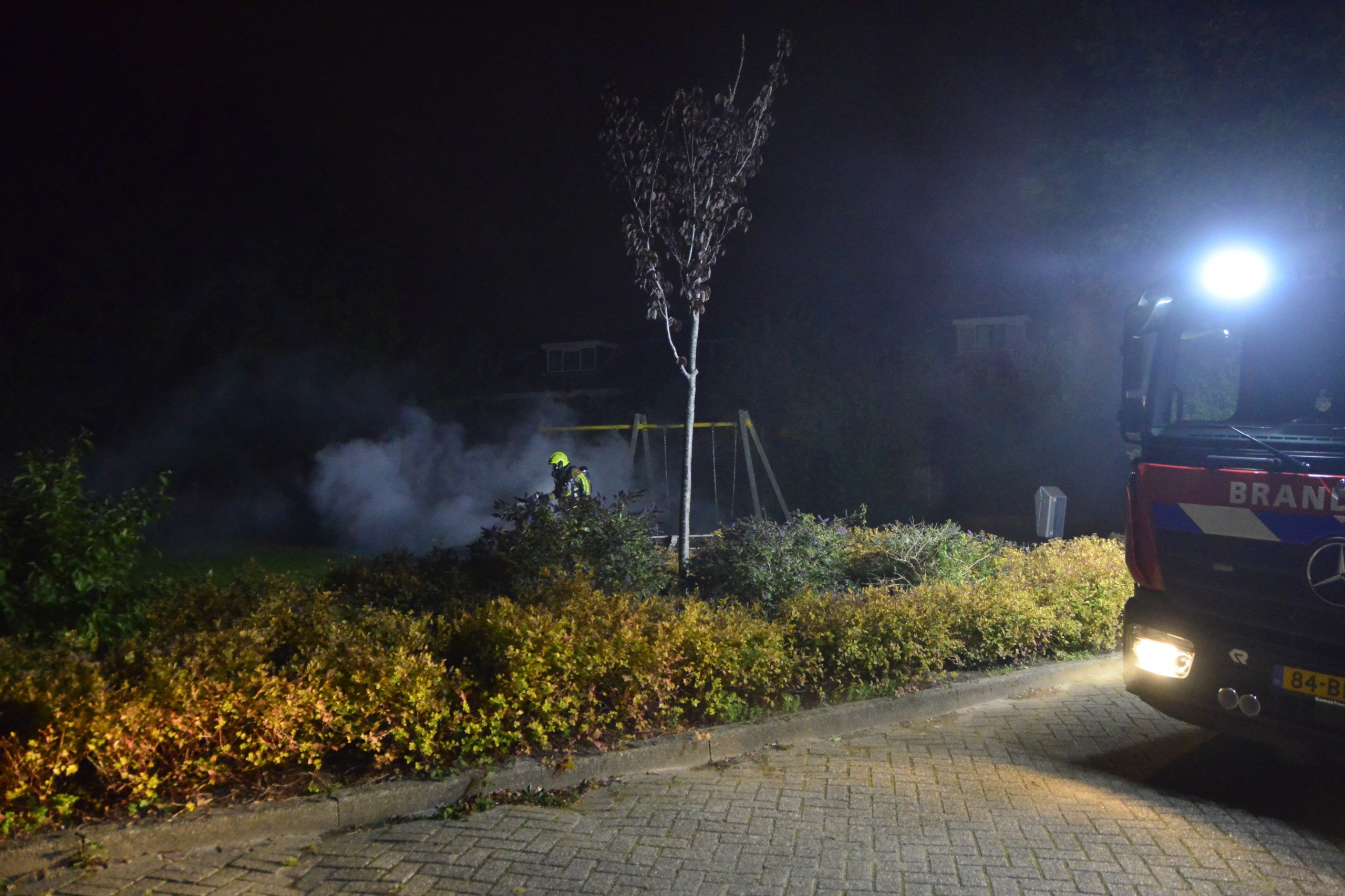 Brandweerman blust brand in een speeltuin 's nachts, brandweerwagen op de voorgrond.