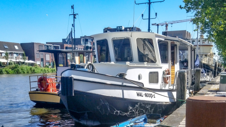 Een wit en zwart schip genaamd "WAL-NOOD1" aangemeerd aan een kade, met moderne gebouwen op de achtergrond en een heldere blauwe lucht.