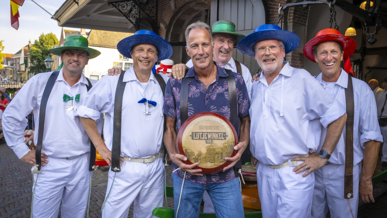 Een groep mannen poseert lachend voor de camera, waarbij één man een kaaswiel vasthoudt. Ze hebben traditionele witte kleding en gekleurde hoeden aan.