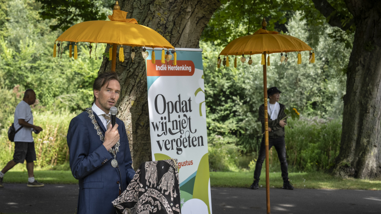 Man houdt een toespraak tijdens de Indië Herdenking, met een bord dat zegt "Opdat wij niet vergeten" en gele parasols op de achtergrond.