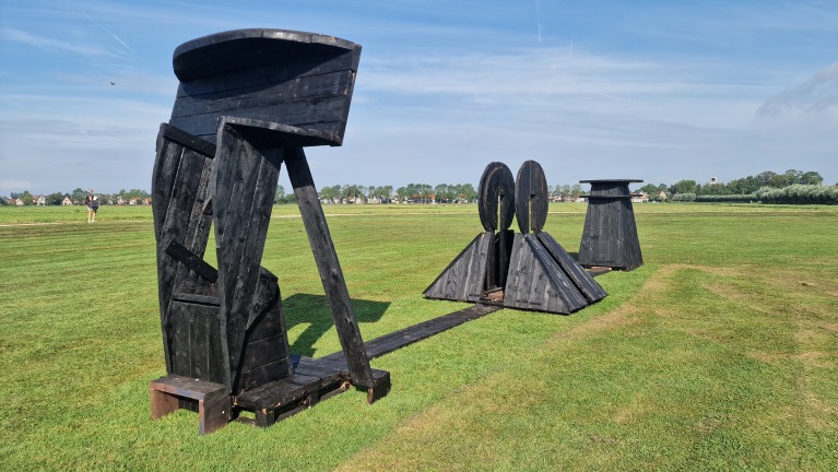 Zwart houten kunstinstallatie op een grasveld met een dorp op de achtergrond.