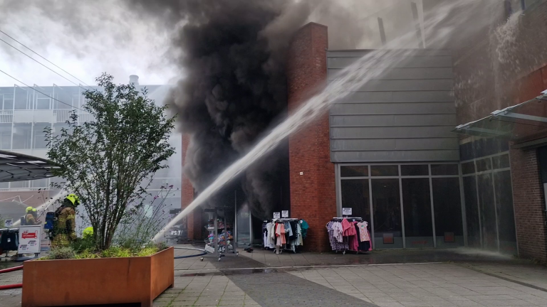 Brandweerlieden blussen een brand in een gebouw, met dikke zwarte rook die uit de ingang komt. Kledingrekken staan buiten de winkel.