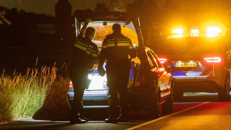 Twee politieagenten controleren een auto langs een donkere weg, verlicht door oranje zwaailichten van een politiewagen.