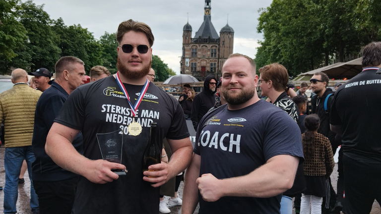 Twee mannen poseren met een trofee en medaille tijdens het evenement "Sterkste Man van Nederland", omringd door andere toeschouwers op een plein met een historisch gebouw op de achtergrond.