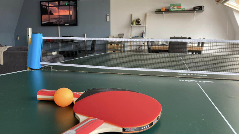 Tafeltennistafel met batjes en een bal, in een woonkamer met een tv op de achtergrond.