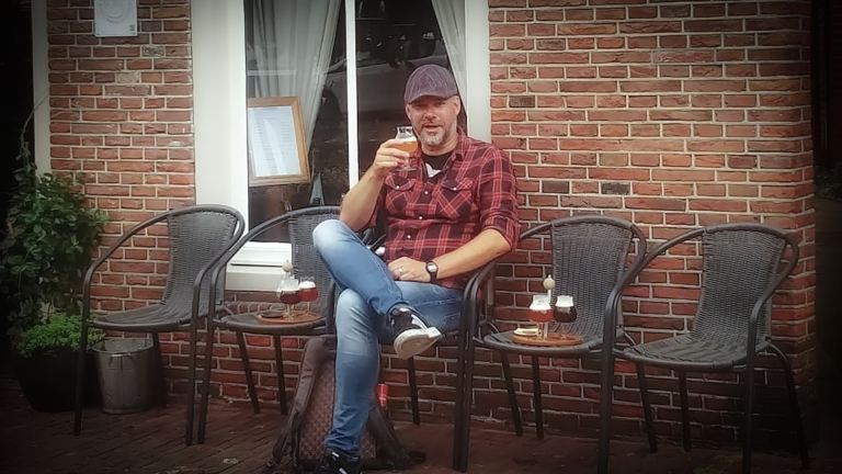 Man in een rood geruit overhemd drinkt bier terwijl hij buiten zit bij een café met bakstenen muren, naast lege stoelen met drankjes erop.