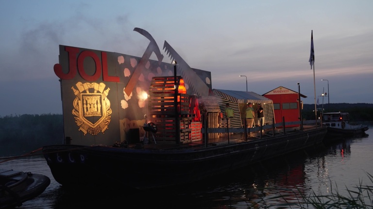 Versierde boot met groot hamer- en zaagsymbool en een bord met de tekst "JOL", mensen op het dek bij schemering.