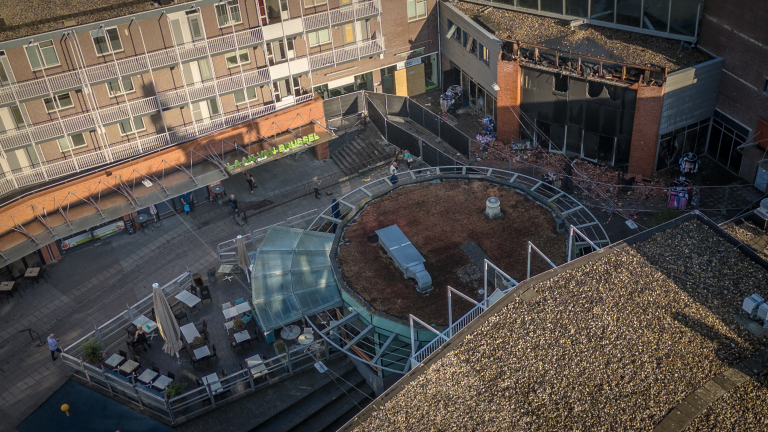 Luchtfoto van een plein omringd door gebouwen, met een overdekte zitgelegenheid en een afgezet gebied met puin van een verwoest gebouw.