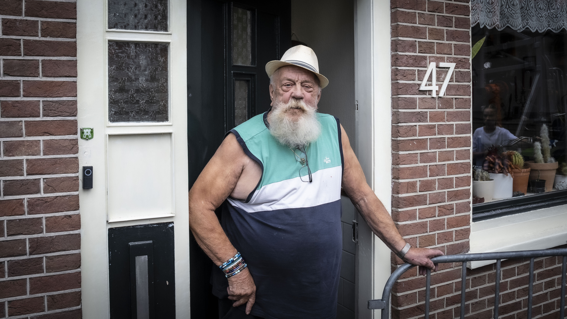 Oudere man met grote witte baard, hoed, en mouwloos shirt staat in de deuropening van een bakstenen huis met het nummer 47.