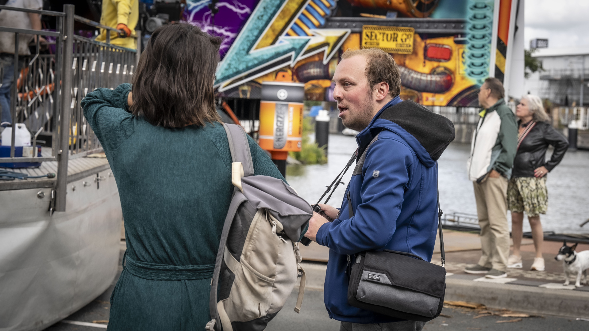 Mensen praten bij een kermisattractie naast een waterkant, met kleurrijke graffiti op de achtergrond.