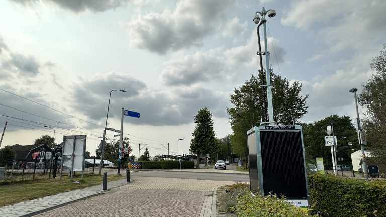 Straat hoek met wegwijzerbord, een parkeerplaats en een beveiligingscamera-installatie, onder een bewolkte hemel.