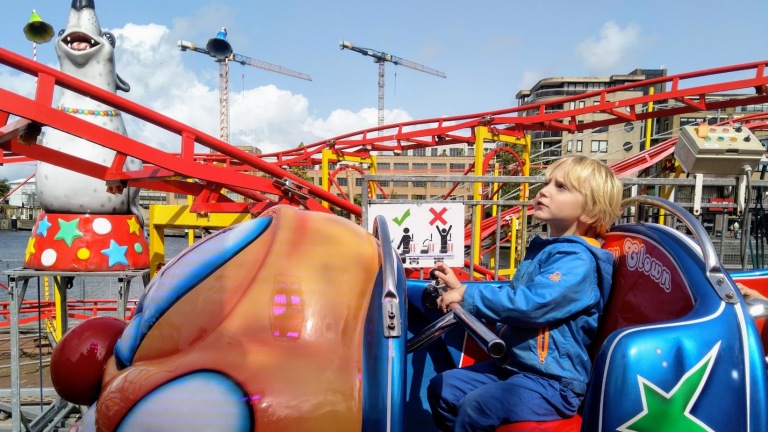 Een jongen in een blauwe jas zit in een achtbaanauto naast een kleurrijke clown-zeehondfiguur.