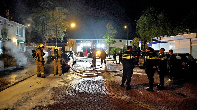 Brandweerlieden en politieagenten bij een nachtelijke autobrand in een woonwijk.