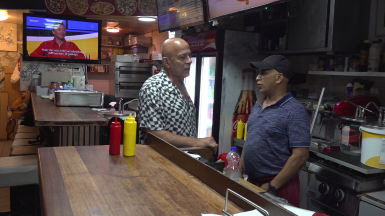 Twee mannen praten in een fastfoodrestaurant met een tv op de achtergrond.