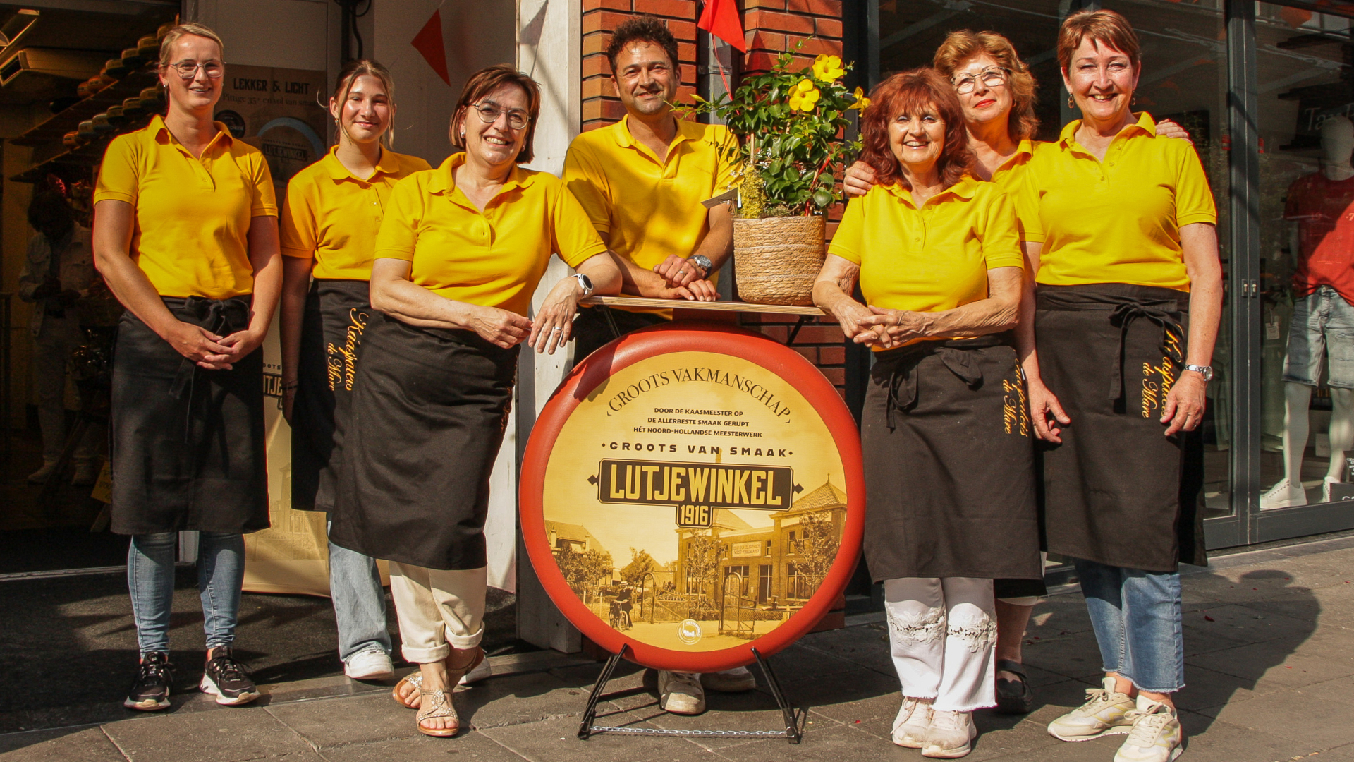 Groepsfoto van zeven medewerkers in gele shirts en zwarte schorten, staand voor een winkel met een groot kaaswiel waarop "Lutjewinkel 1916" staat.