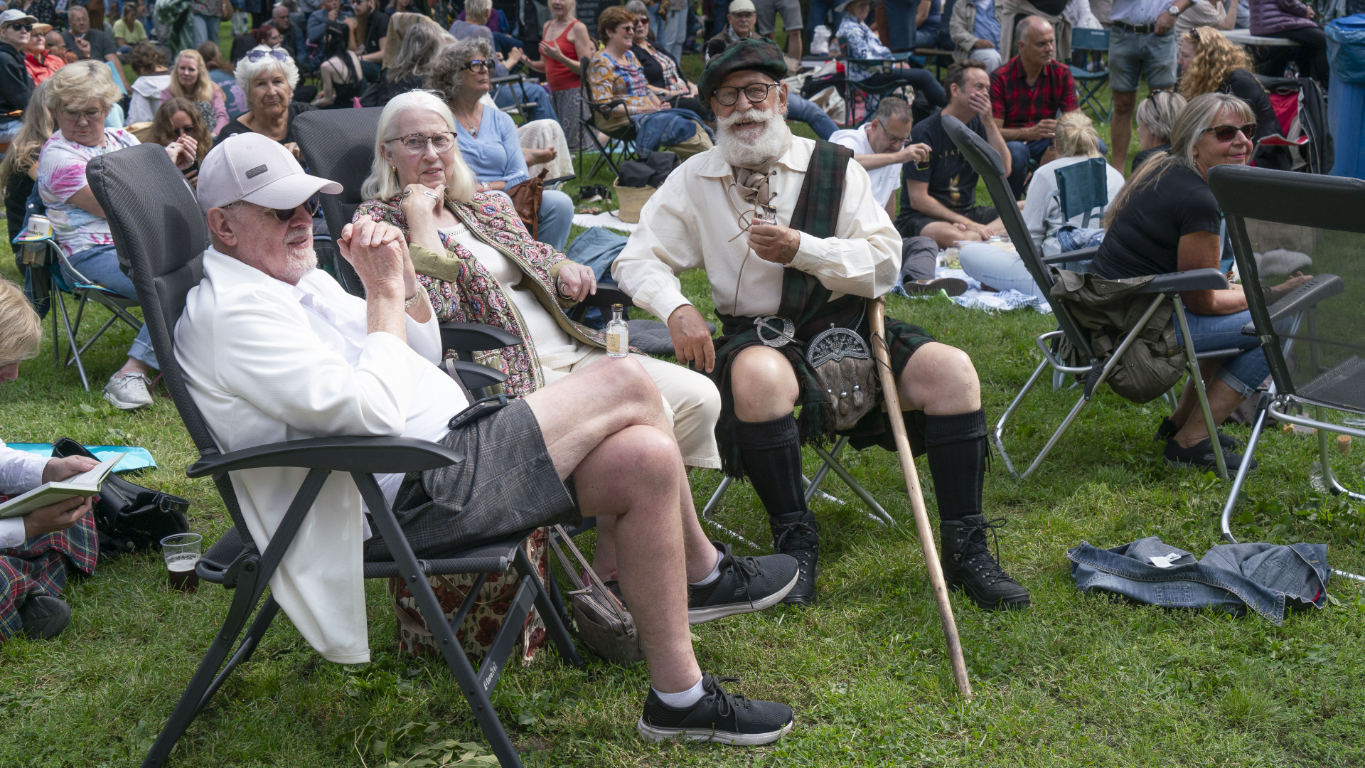 Een groep oudere mensen zit buiten op stoelen tijdens een festival, waarbij een man traditionele Schotse kleding draagt.