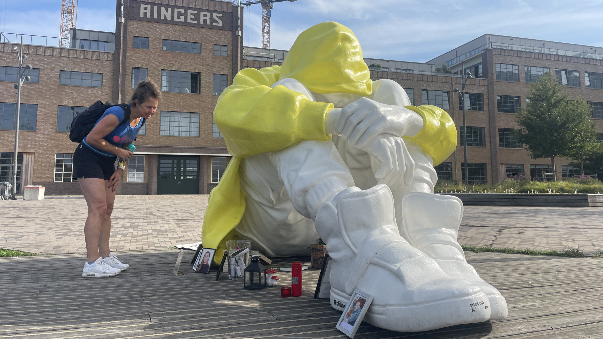 Vrouw kijkt naar groot wit-gele standbeeld van een figuur met een capuchon en opgeheven knieën voor een historisch gebouw met het woord "Ringers".