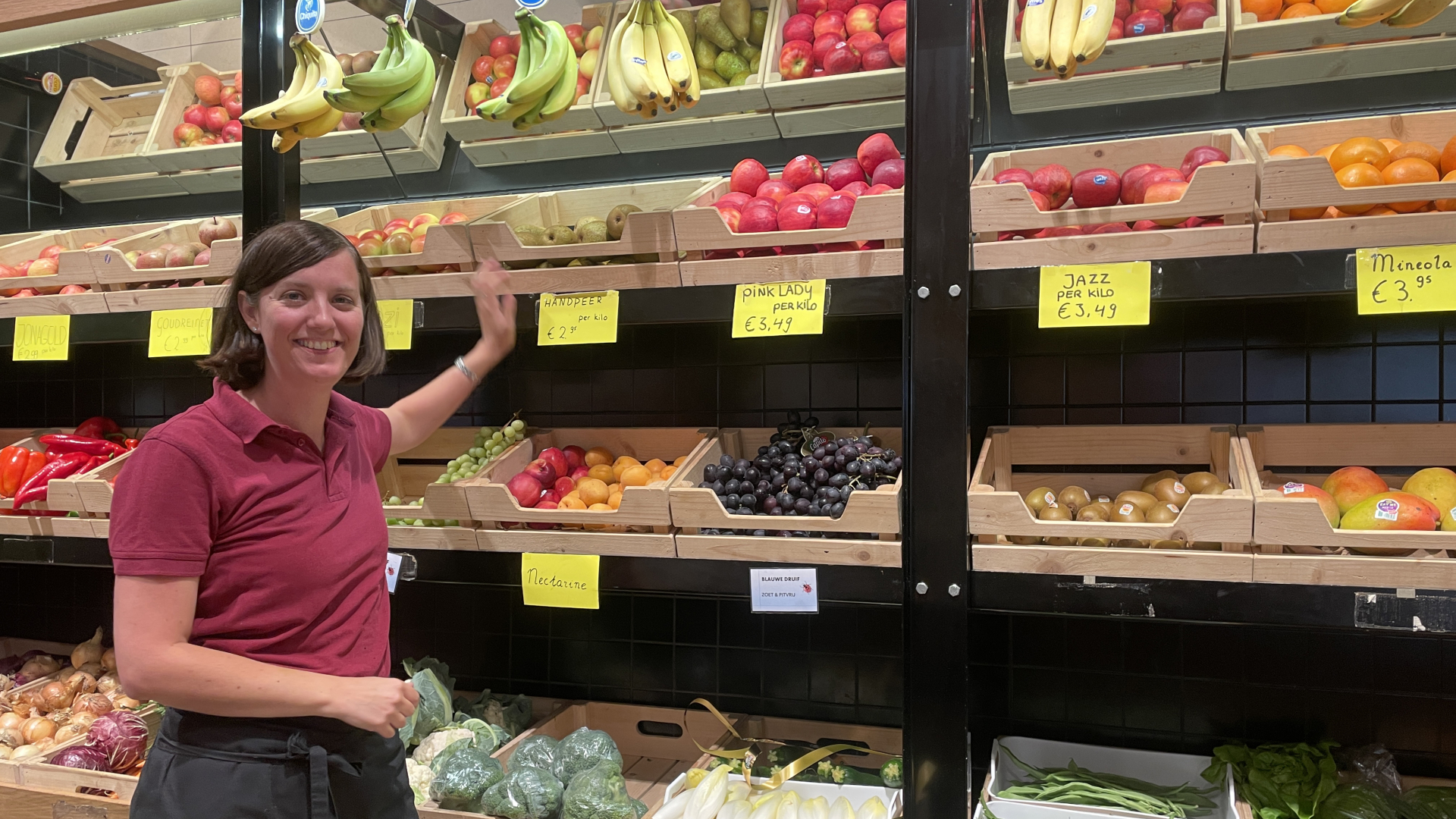 Medewerkster in roodgeklede polo arrangeert fruit en groente in een supermarkt.