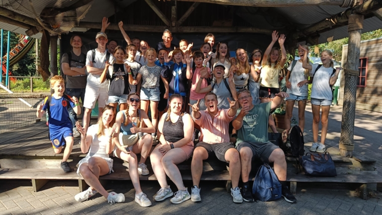 Groep mensen poseren enthousiast op een houten podium in een buitenomgeving.