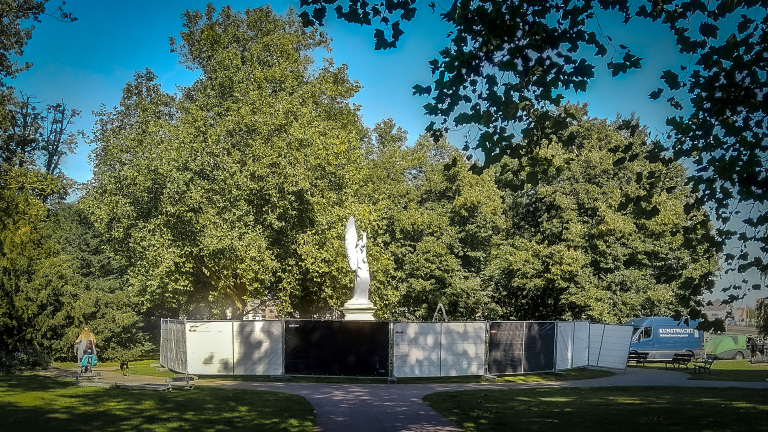 Beeldhouwwerk omgeven door bouwhekken in een park met veel bomen.