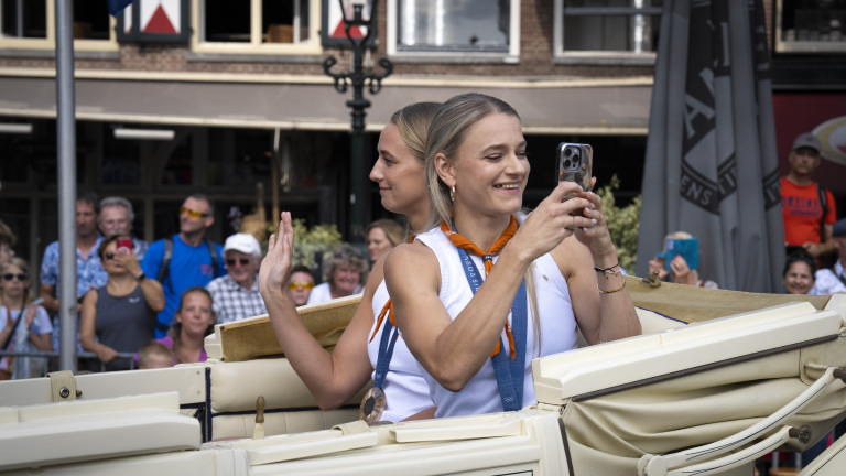 Twee jonge vrouwen maken foto's en zwaaien naar de menigte tijdens een parade in een open voertuig.