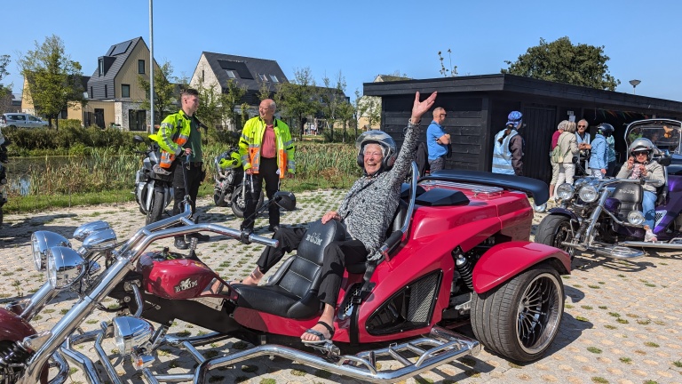 Man zit lachend in een rode driewieler met opgeheven hand, omringd door mensen en andere motoren op een zonnige dag.