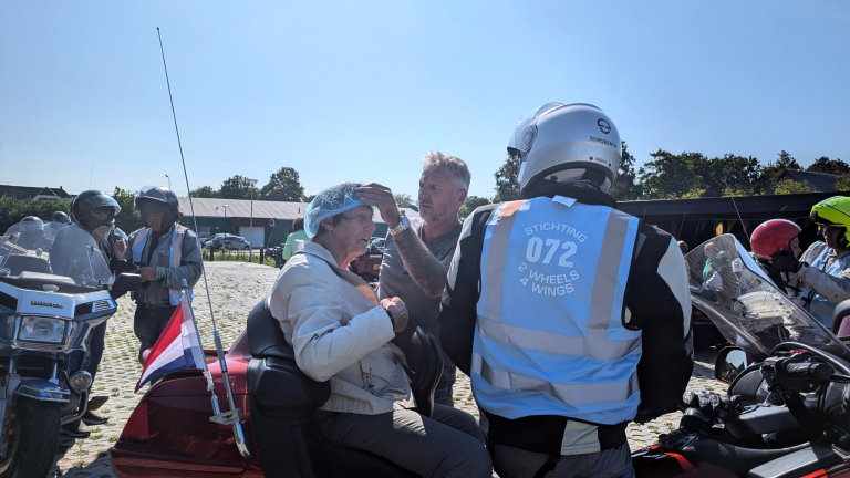 Een oudere vrouw zit op een motorfiets terwijl een man haar helpt met een haarnetje. Achter hen staat een man met een reflecterend vest met de tekst "Stichting 072 2 Wheels 4 Wings". Andere mensen en motorfietsen zijn op de achtergrond zichtbaar.