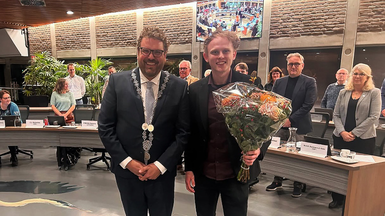 Twee mannen staan lachend in een zaal, één houdt een boeket bloemen vast. Op de achtergrond zijn mensen en een display te zien.