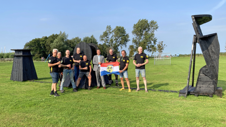 Een groep mannen, gekleed in zwarte shirts en korte broeken, poseren buiten op een grasveld naast grote houten sculpturen, waarbij een van hen een vlag vasthoudt.