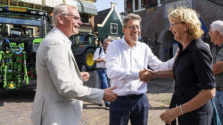 Drie mensen, twee mannen in lichte kleding en een vrouw in een zwarte blouse, schudden elkaar lachend de hand op een plein met een tractor op de achtergrond.