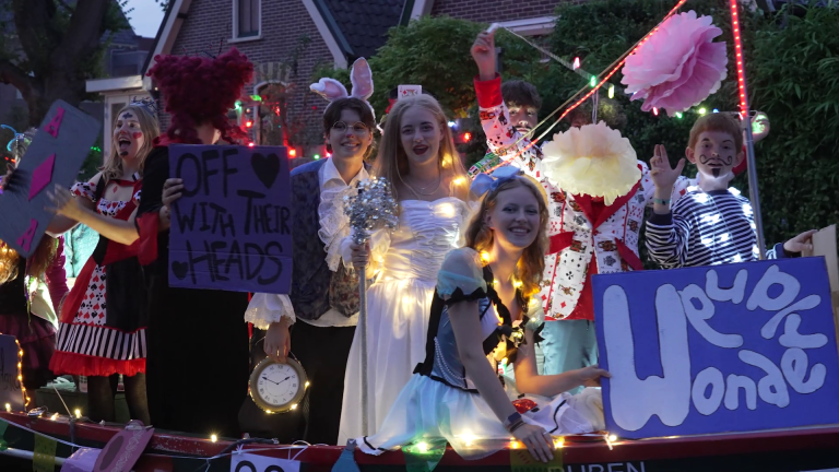 Groep mensen verkleed als personages uit "Alice in Wonderland" staan op een versierde wagen met lichtjes en borden.