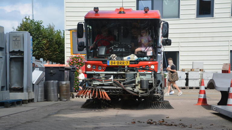 Rode straatveegmachine met kinderen erin, geparkeerd naast afvalcontainers en kegels, met gebouw op de achtergrond.